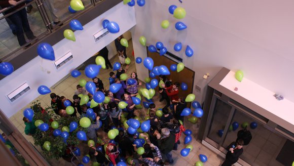 Green and blue balloons rise to the ceiling as students look up at them.