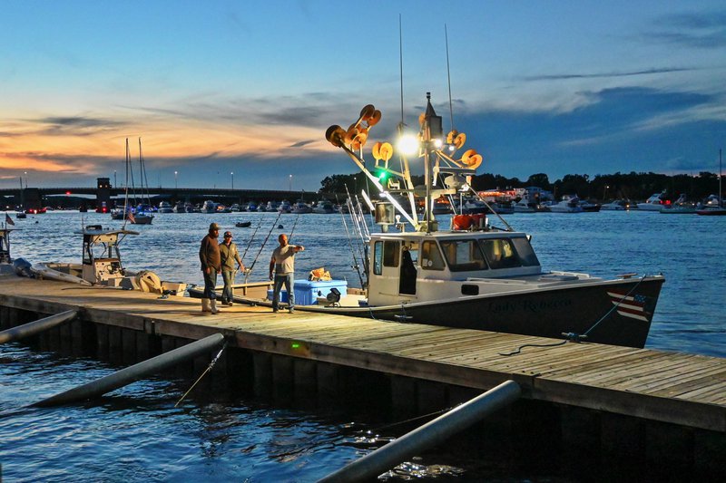 A fisherman directs his crew on a dock, next to a fishing boat in the twilight.