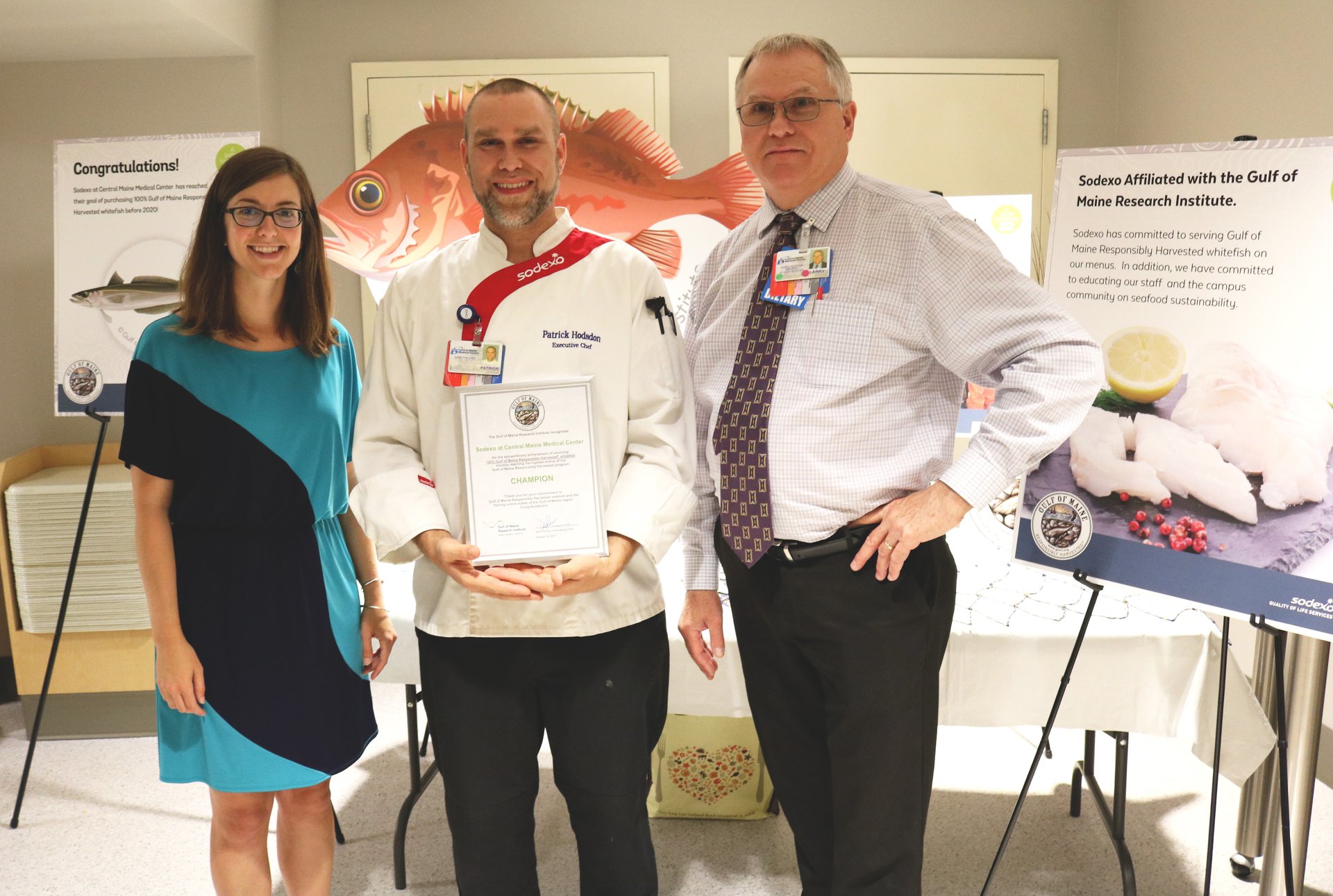 Kyle Foley, our sustainable seafood program manager, stands in a blue and black dress next to the right of to two men as they pose for a photo in celebration of the hospital shifting to all local seafood. The man in the middle holds a certificate acknowledging the achievement.