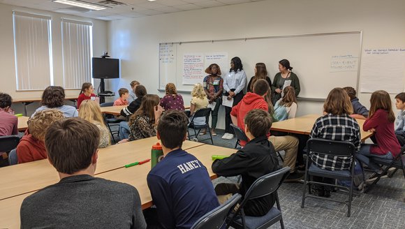 Students sit in a classroom and listen to other students presenting at the front of the class.