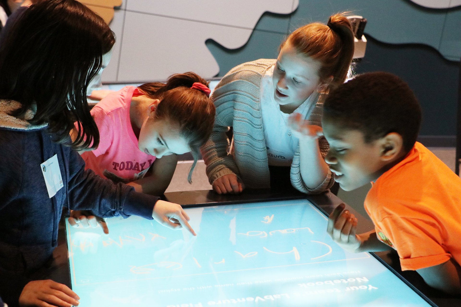 Four students are gathered around a large, flat touch table as they talk about what is happening on it.
