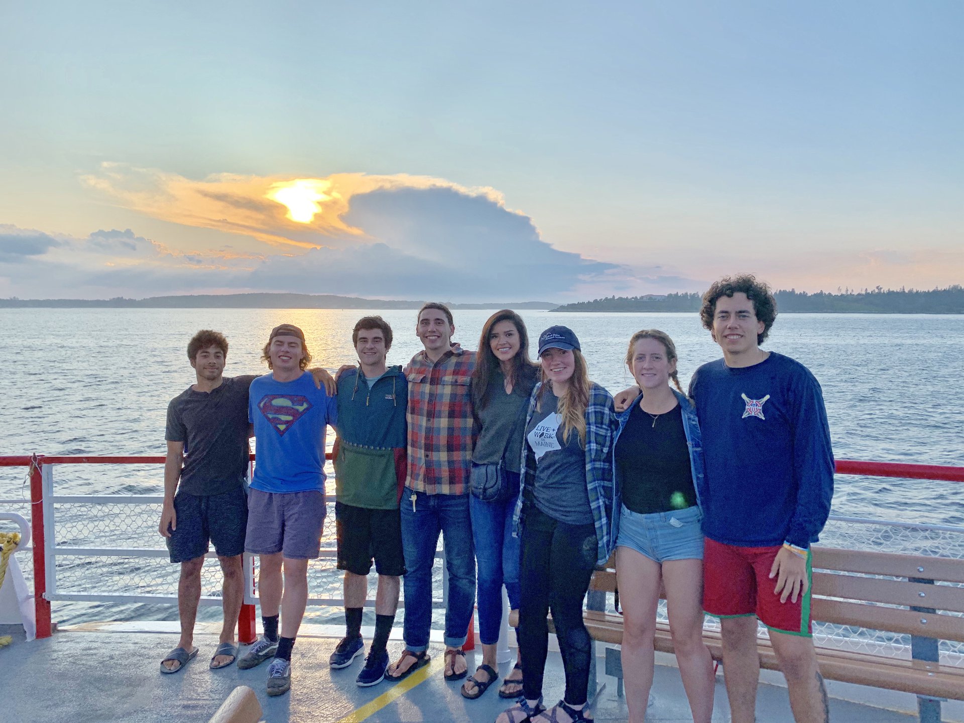 Eight REU students pose smiling for a photo along a boat's railing with the sun setting
over the water behind them.
