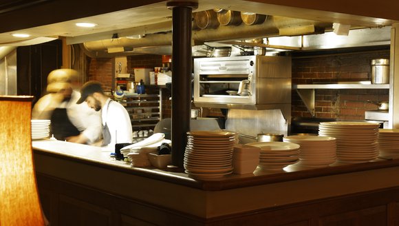Chefs working in a open restaurant kitchen with stacks of plates in the foreground