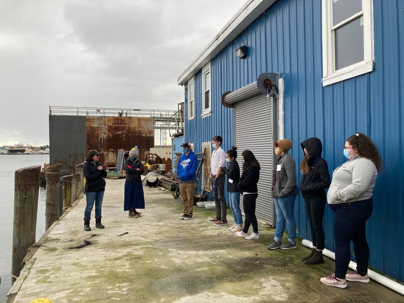 10 students stand in a line on the right side of the photo, facing a speaker who is on the left side of the photo. They are standing on a concrete dock on a wharf, with a blue building behind them.