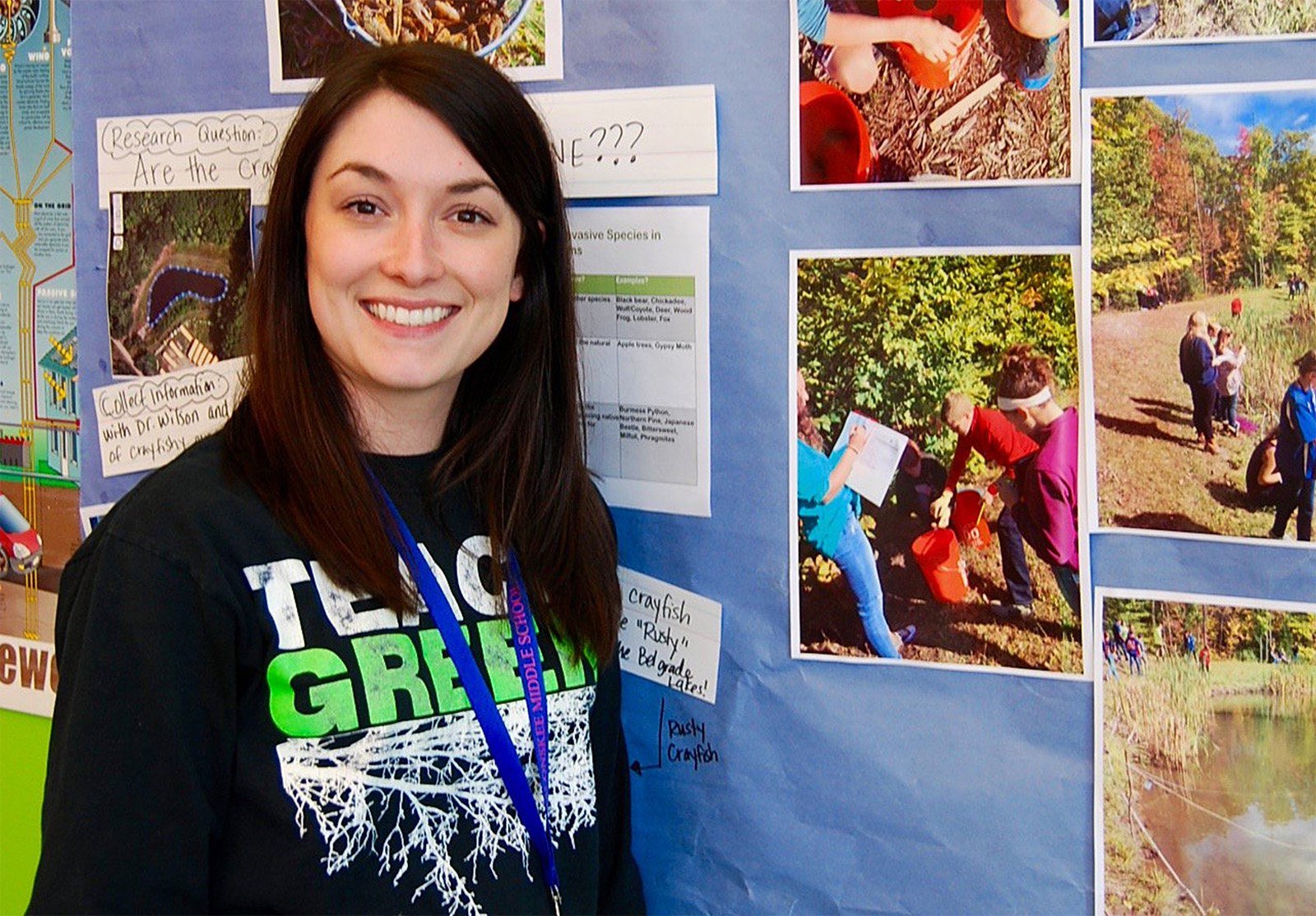 Messalonskee Middle School teacher Amanda Ripa stands in a black shirt with a blue lanyard on in front of a wall plastered with her student's work on invasive species.