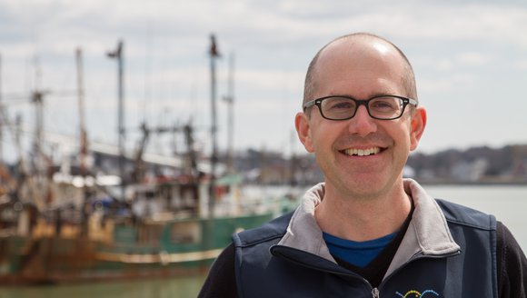Chief Scientific Officer Andy Pershing poses outside in the sunlight for a portrait in front of a marina, wearing a blue vest with Gulf of Maine Research Institute written on the upper left.