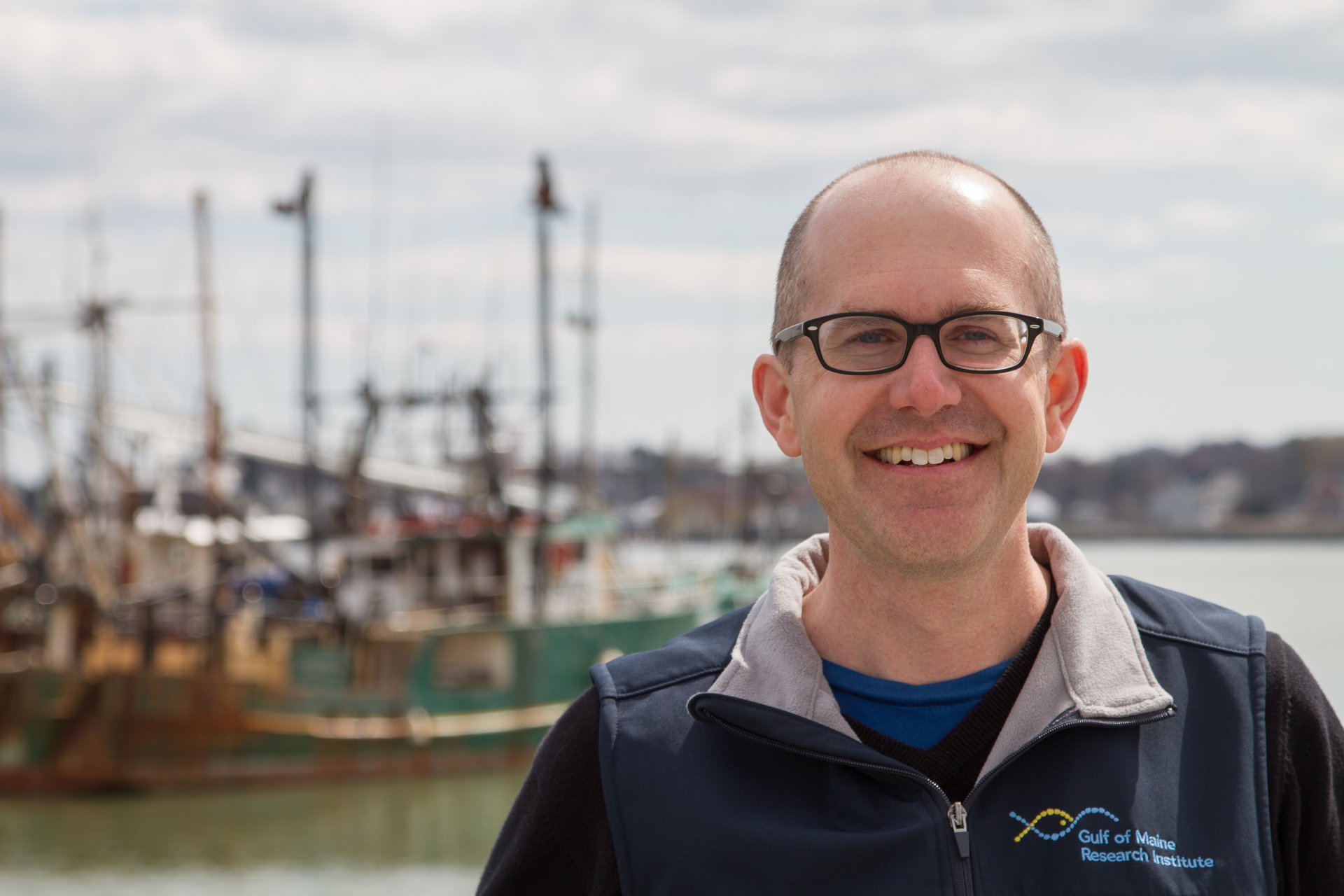 Chief Scientific Officer Andy Pershing poses outside in the sunlight for a portrait in front of a marina, wearing a blue vest with Gulf of Maine Research Institute written on the upper left.