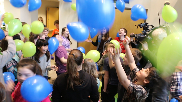 Dozens of joyful students and teachers laugh and smile in a room filled with green and blue balloons.