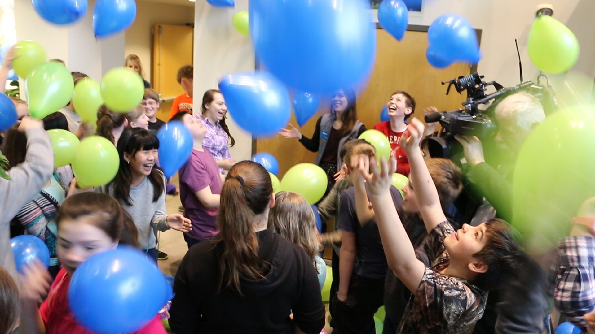 Dozens of joyful students and teachers laugh and smile in a room filled with green and blue balloons.