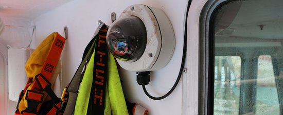 A camera with a round lens and a white circular base sits mounted on the wall of a ship.