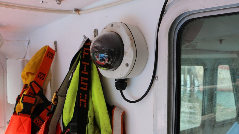 A camera with a round lens and a white circular base sits mounted on the wall of a ship.