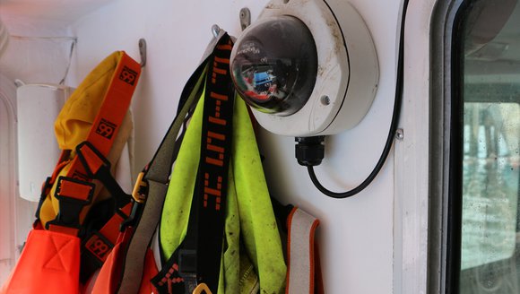 A camera with a round lens and a white circular base sits mounted on the wall of a ship.