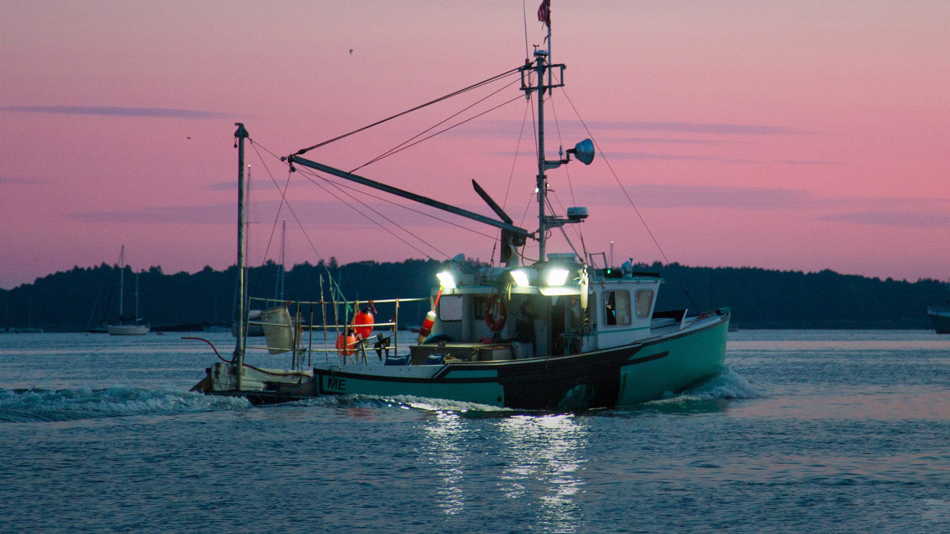 A fishing vessel motors to the right in front of a pink sunset sky.