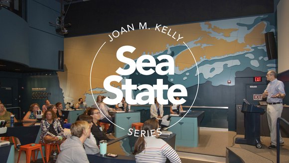 Behind a diagram in white that reads "Joan M. Kelly Sea State Series" participants sitting on orange stools engage in our Sea State Series by listening to a GMRI expert who stands on a platform to the right of the image. The event took place in the LabVenture space, and the big brown, blue, and black map with the Gulf of Maine sits in the background.