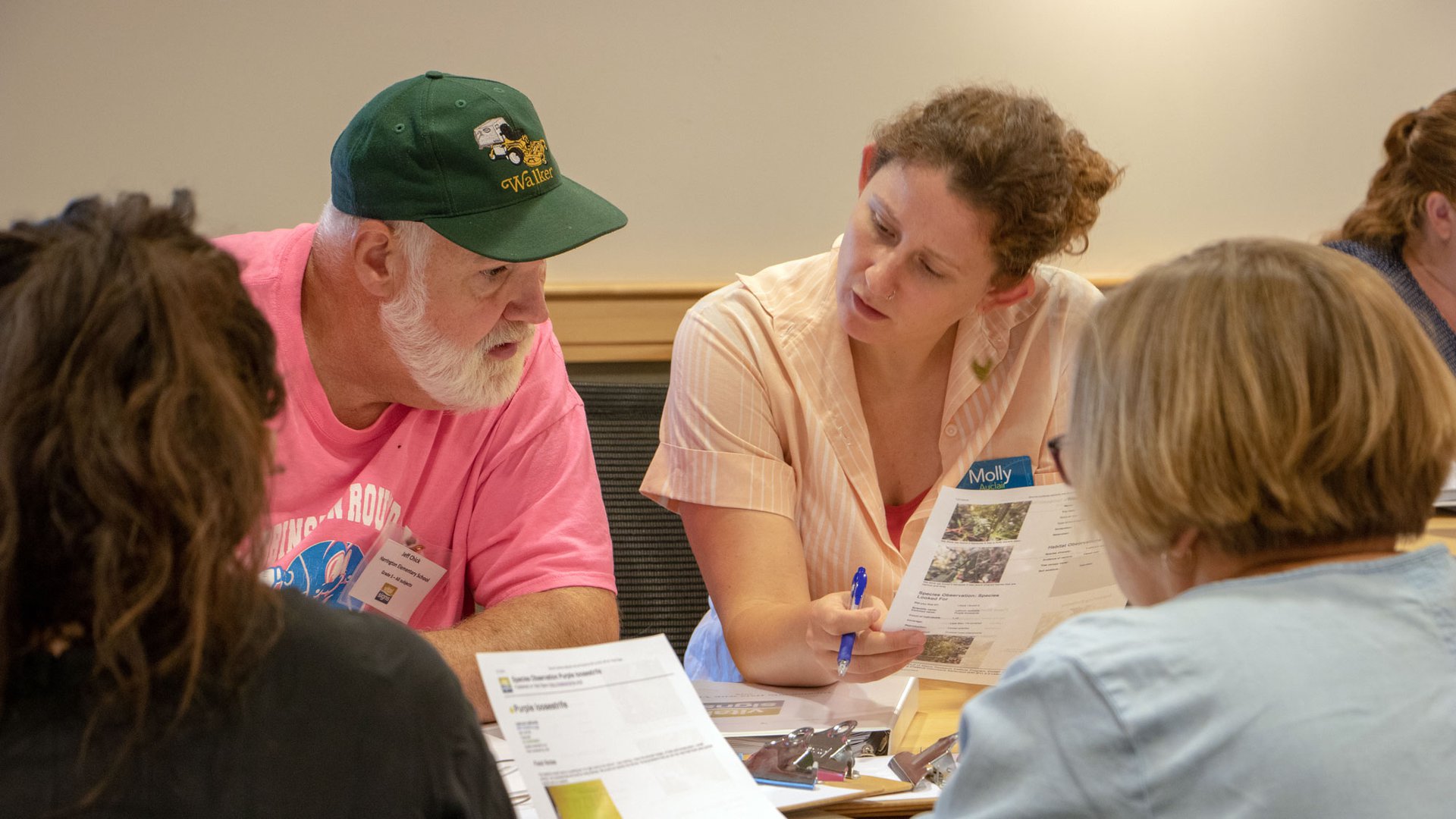 Maine teachers — one a man in a green hat and a bright pink shirt, and the other a woman in a salmon colored shirt — converse at light brown table at a teacher workshop.