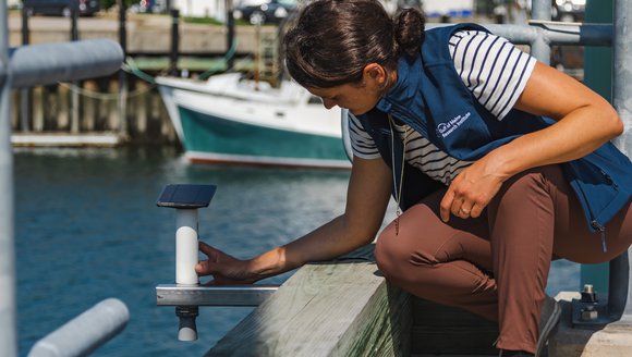 A woman in a blue vest and brown pants kneels down next to a rectangular, black device hanging out over a pier.