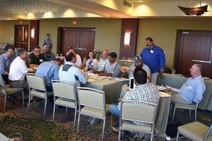 People are sitting at a long table in a large carpeted room.