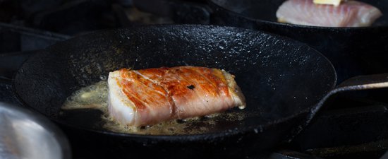 A rectangular piece of fish is browning on a black frying pan in the foreground, while a more raw piece of fish with a square of butter melts on top.