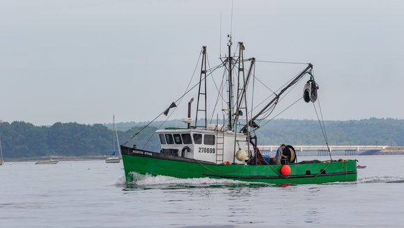 A green fishing boat is heading toward the left side of the photo on smooth blue waters. A smaller boat is blurred in the distance.