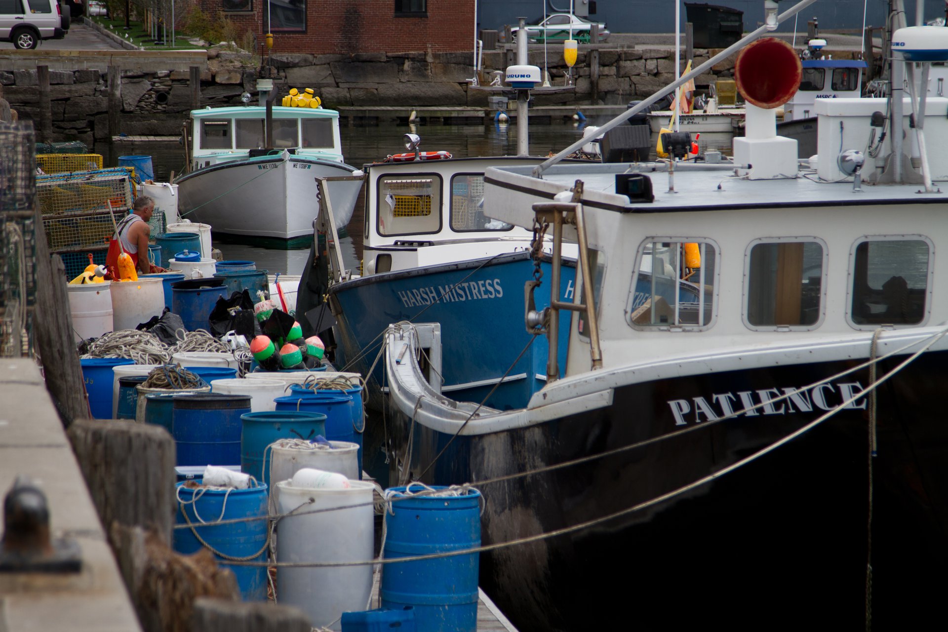 Three lobster boats sit next to a dock facing the camera, loaded with different colored lobster traps. The dock is on the left of the photo, and the boats are on the right. The boat in the foreground is black, the middle boat is teal, and the boat in the back is white. A man in orange waders weaves through the lobster traps on the dock and is walking toward the middle boat.